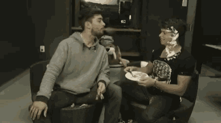 a man is blowing smoke into another man 's face while sitting on a couch with a plate of food .