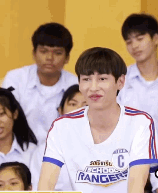 a young man wearing a school rangers shirt is sitting in a classroom with other students .