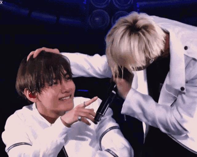 a man in a white jacket is touching another man 's head while holding a microphone