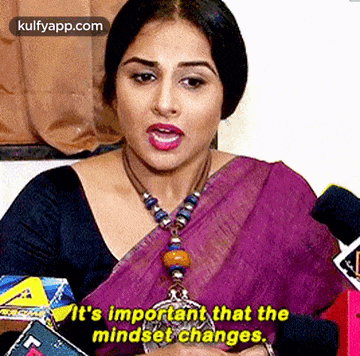a woman in a purple saree is sitting in front of a microphone and talking .