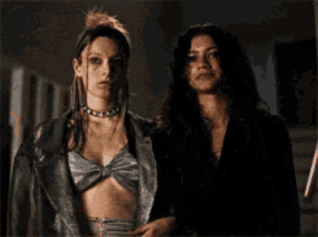 two women are standing next to each other in a dark room . one of the women is wearing a leather jacket .