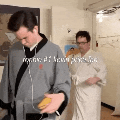 two men in bathrobes are standing next to each other in a room . one of the men is listening to music on his phone .