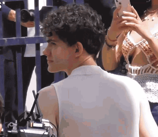 a man with curly hair is standing in front of a fence while a woman takes a picture of him