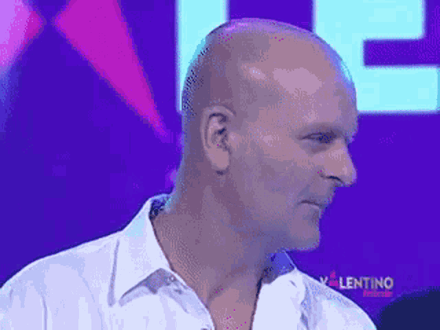 a bald man in a white shirt is sitting in front of a purple background with the word valentino on it .