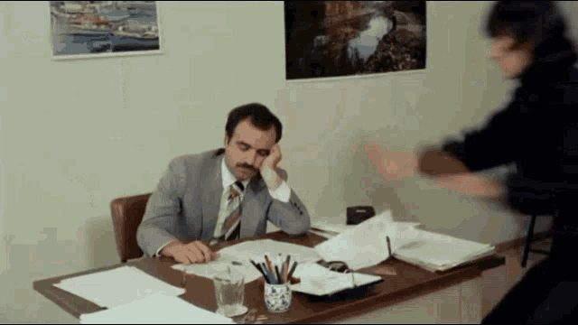 a man in a suit and tie is sitting at a desk with papers and a glass of water