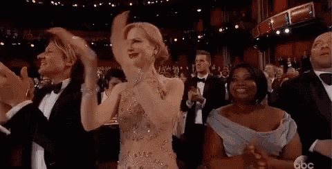 a woman in a dress is applauding while standing in front of a crowd of people in tuxedos .