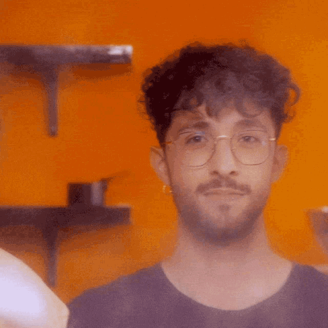 a man with glasses and a beard looks at the camera with an orange background