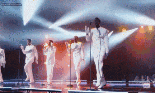 a group of men in white suits are singing into microphones on stage .