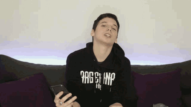 a young man sitting on a couch holding a cell phone and wearing a hoodie that says ' rage '