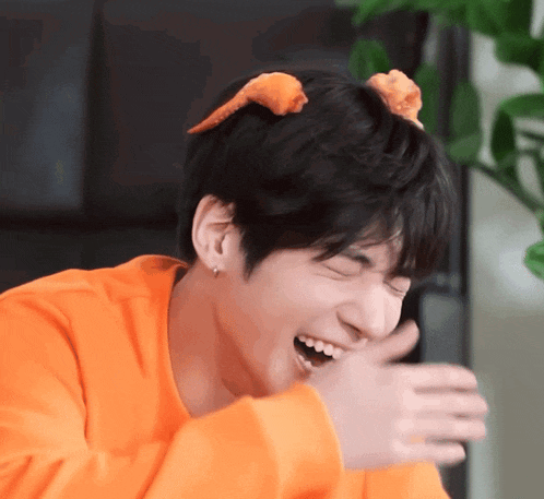 a young man wearing an orange sweater is laughing and covering his mouth .
