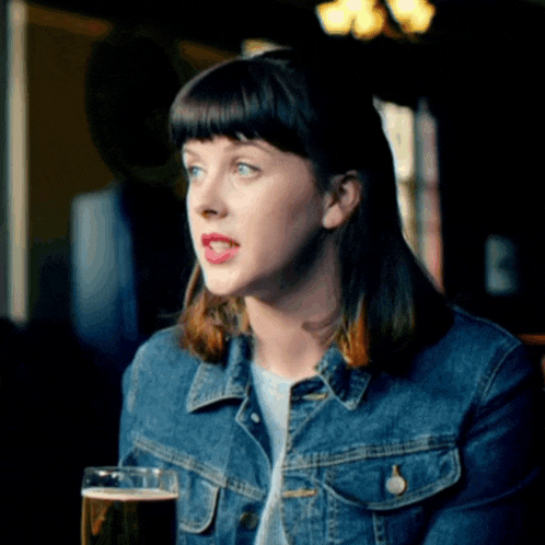 a woman in a denim jacket holds a glass of beer in her hand