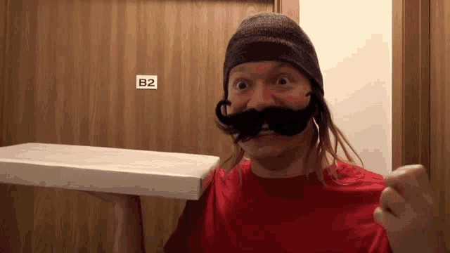 a man with a fake mustache is holding a pizza box in front of a door that says b2 on it