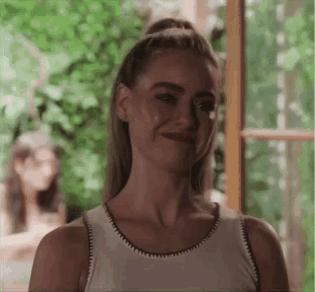 a woman in a white tank top is standing in front of a window .