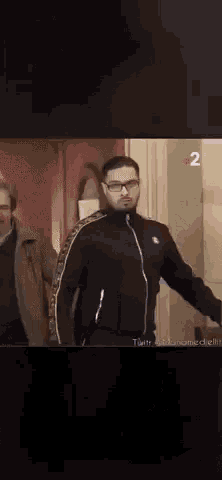 a man wearing glasses and a black jacket is walking down a hallway with two other men .