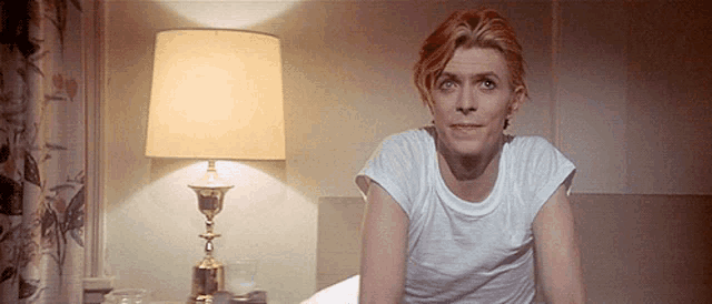 a man in a white shirt is sitting on a bed in front of a lamp