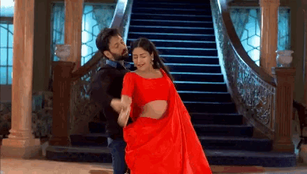 a man and a woman are dancing in front of stairs . the woman is wearing a red saree .