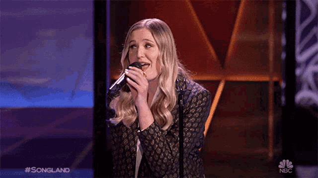 a woman singing into a microphone on a stage with the nbc logo behind her