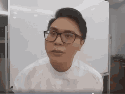 a man wearing glasses and a white shirt is sitting in front of a whiteboard .