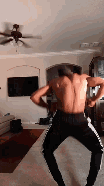 a shirtless man is dancing in a living room with a ceiling fan in the background