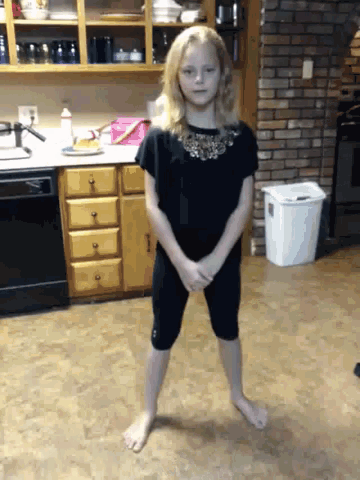 a little girl is standing in a kitchen with a trash can in the background
