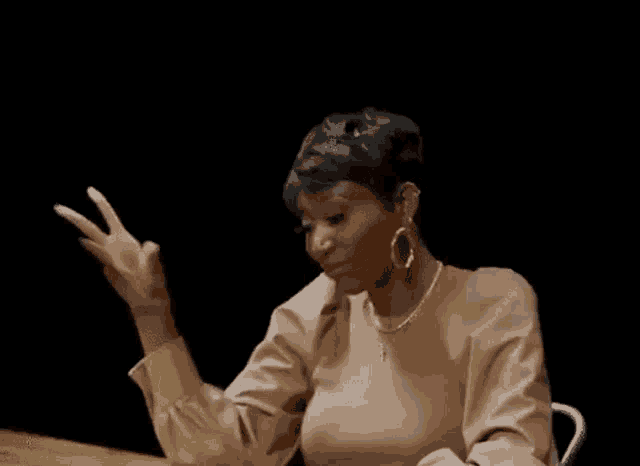 a woman in a brown shirt is sitting at a table with her hand up .