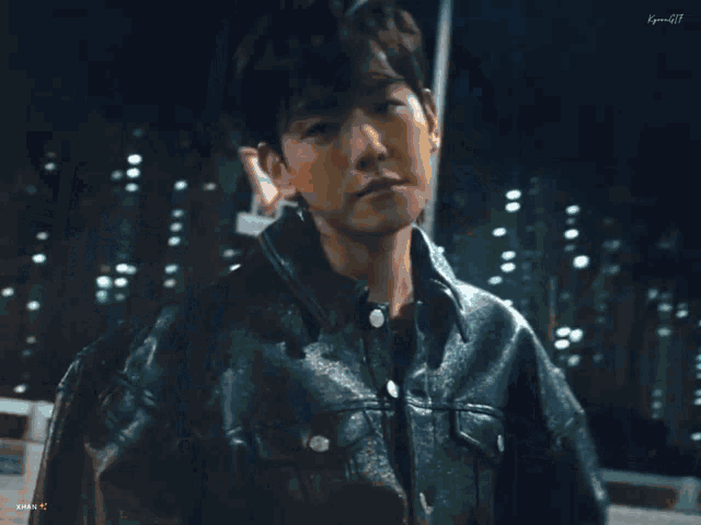 a young man wearing a black leather jacket is standing in front of a city street at night .
