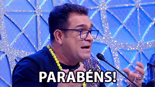a man wearing glasses is speaking into a microphone and the word parabéns is on the screen