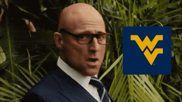 a bald man wearing glasses and a suit stands in front of a blue w logo