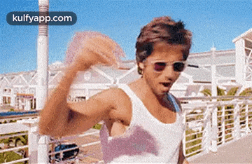 a man in a white tank top and sunglasses is flexing his muscles on a bridge .