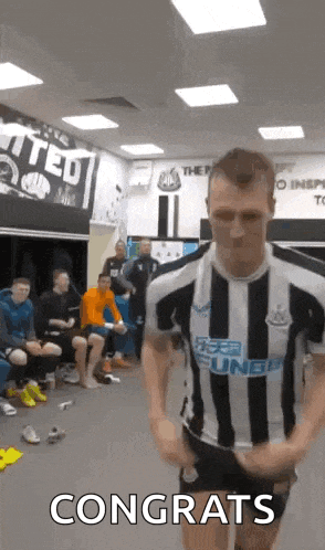 a man in a striped shirt is standing in a locker room with a group of people sitting in the background .