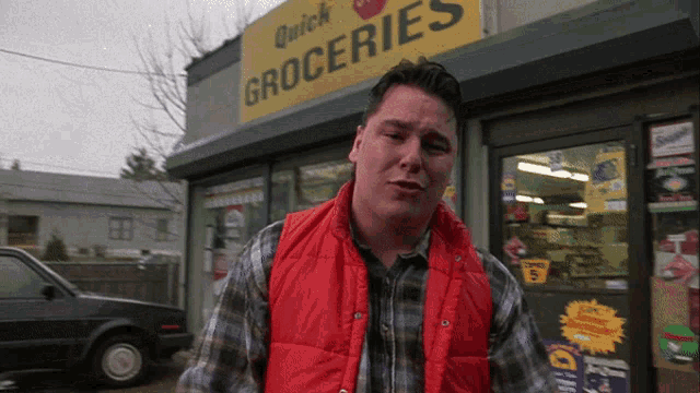 a man wearing a red vest stands in front of a quick groceries store