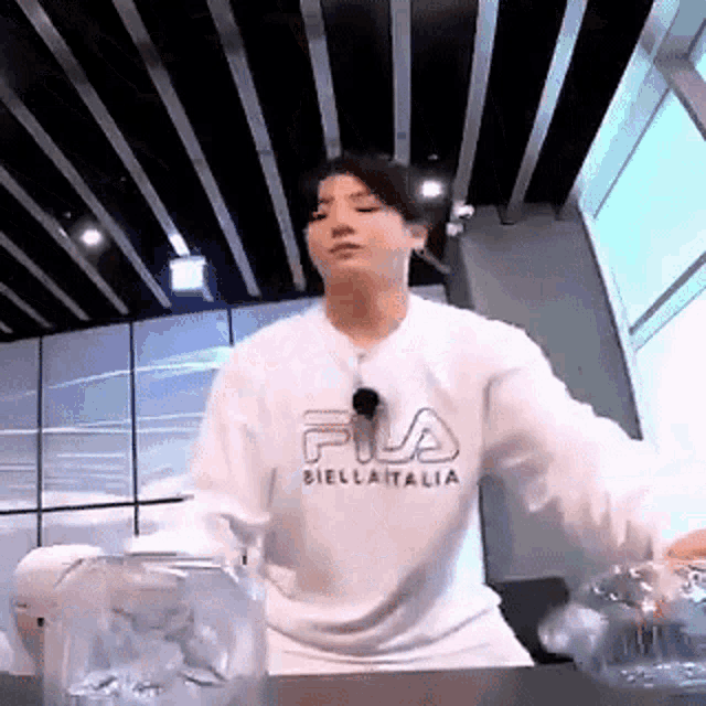 a young man in a white fila sweatshirt is standing in front of a table holding a bottle of water .