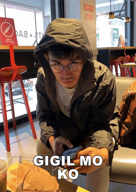 a man wearing a hooded jacket and glasses is looking at his phone with gigil mo ko written on the bottom