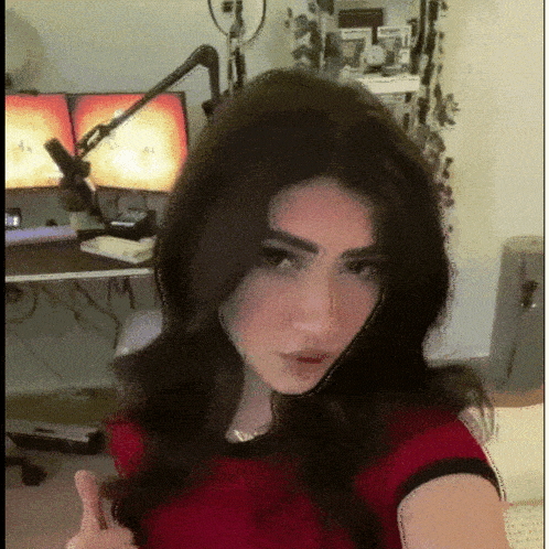 a woman in a red shirt is giving a thumbs up in front of two computer monitors