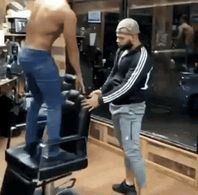 a man is standing on a barber chair in a barber shop while another man holds his hand .
