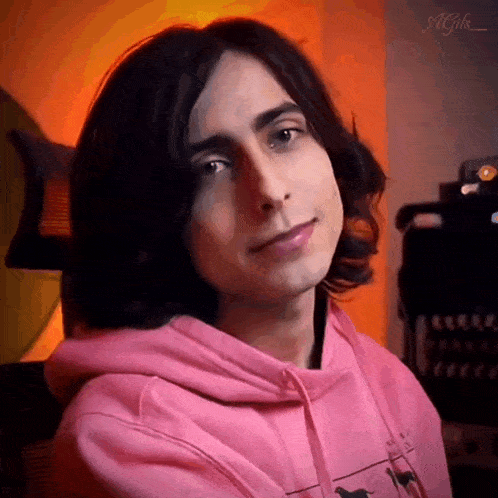 a young man with long dark hair is wearing a pink hoodie and smiling .