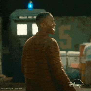 a man in a plaid jacket is standing in front of a disney + sign .