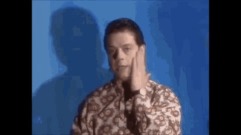 a man in a floral shirt is covering his face with his hand in front of a blue background .