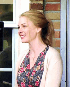 a woman wearing a floral shirt and a tan cardigan smiles in front of a brick wall