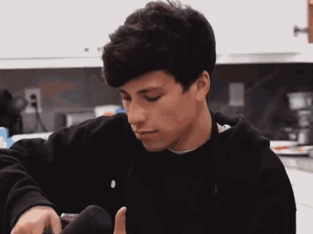 a young man in a black hoodie is sitting in a kitchen looking down at something .