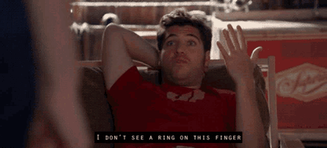 a man in a red shirt is sitting in a chair and says i don 't see a ring on this finger