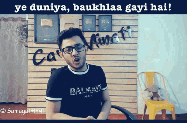 a man wearing glasses and a balmain shirt is standing in front of a wooden wall