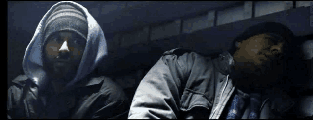 a man wearing a hooded jacket is standing next to another man who is sleeping
