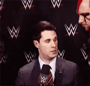 a man in a suit and tie is speaking into a microphone in front of a wall with wrestlers on it