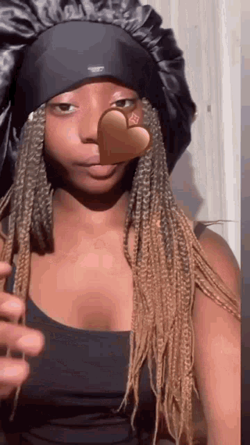 a woman with braids is wearing a black satin hat and holding a heart in front of her face .