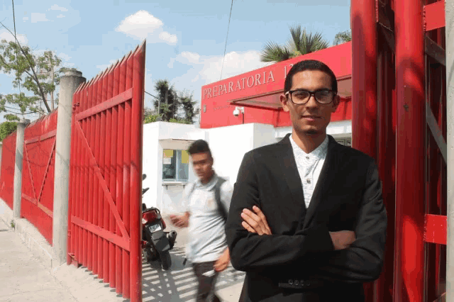 a man in a black suit stands in front of a red fence that says preparatoria