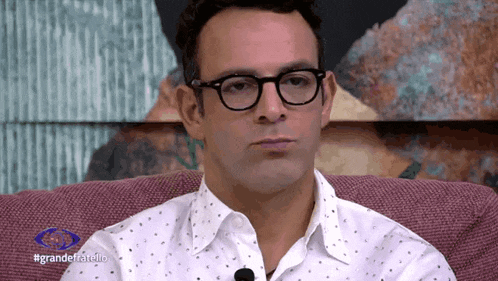 a man wearing glasses and a white shirt is sitting on a couch in front of a sign that says #grandefratello