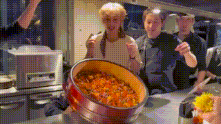 a group of people are standing around a pot of food in a kitchen .