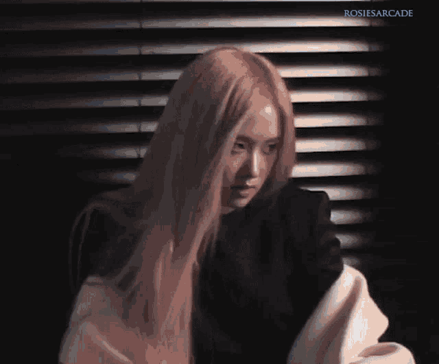 a woman with long pink hair is standing in front of a window with blinds ..