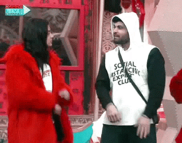 a woman in a red coat stands next to a man wearing a white shirt that says social distancing expert club